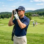 Golfer looking through a black laser rangefinder on a green golf course.